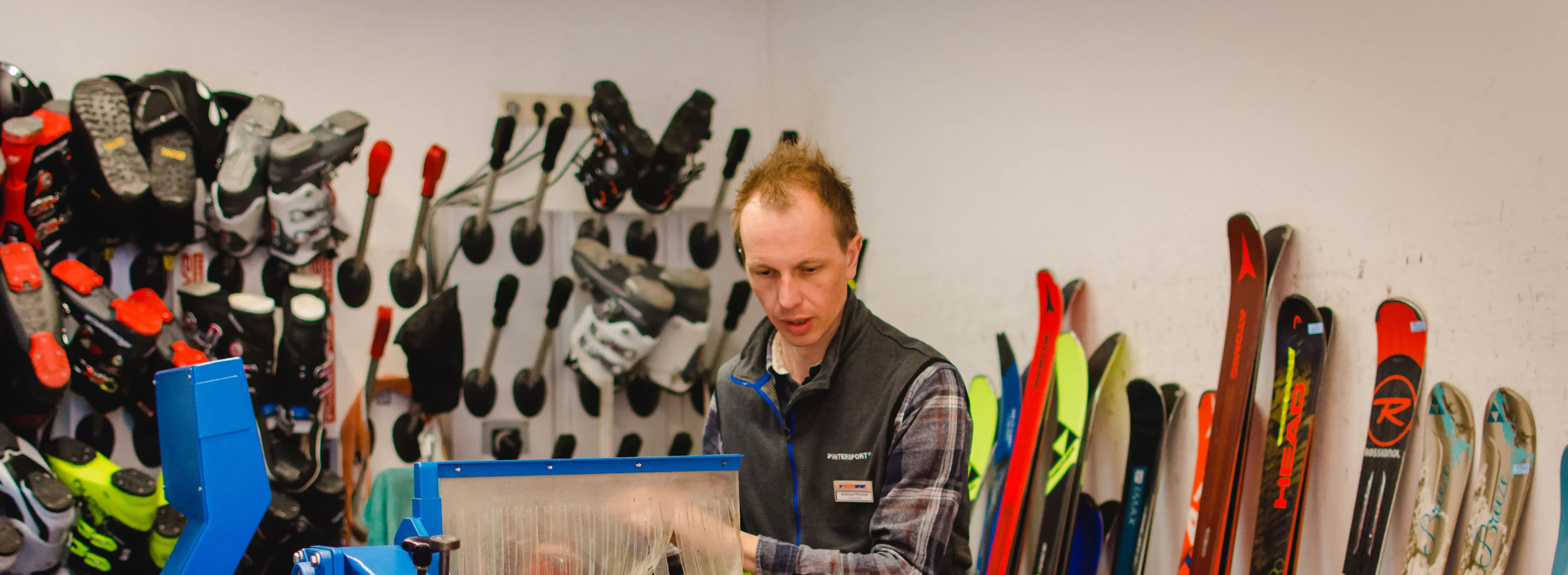A person working in a workshop with ski equipment. Skis and ski boots are lined up on the walls, and the person is concentrating on operating a blue machine.