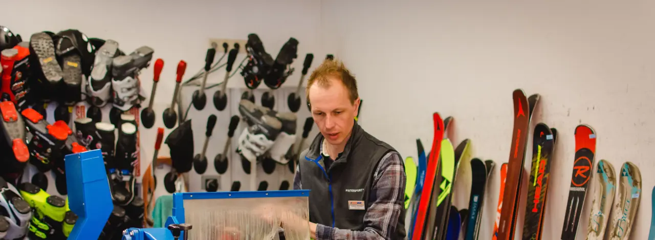 A person working in a workshop with ski equipment. Skis and ski boots are lined up on the walls, and the person is concentrating on operating a blue machine.