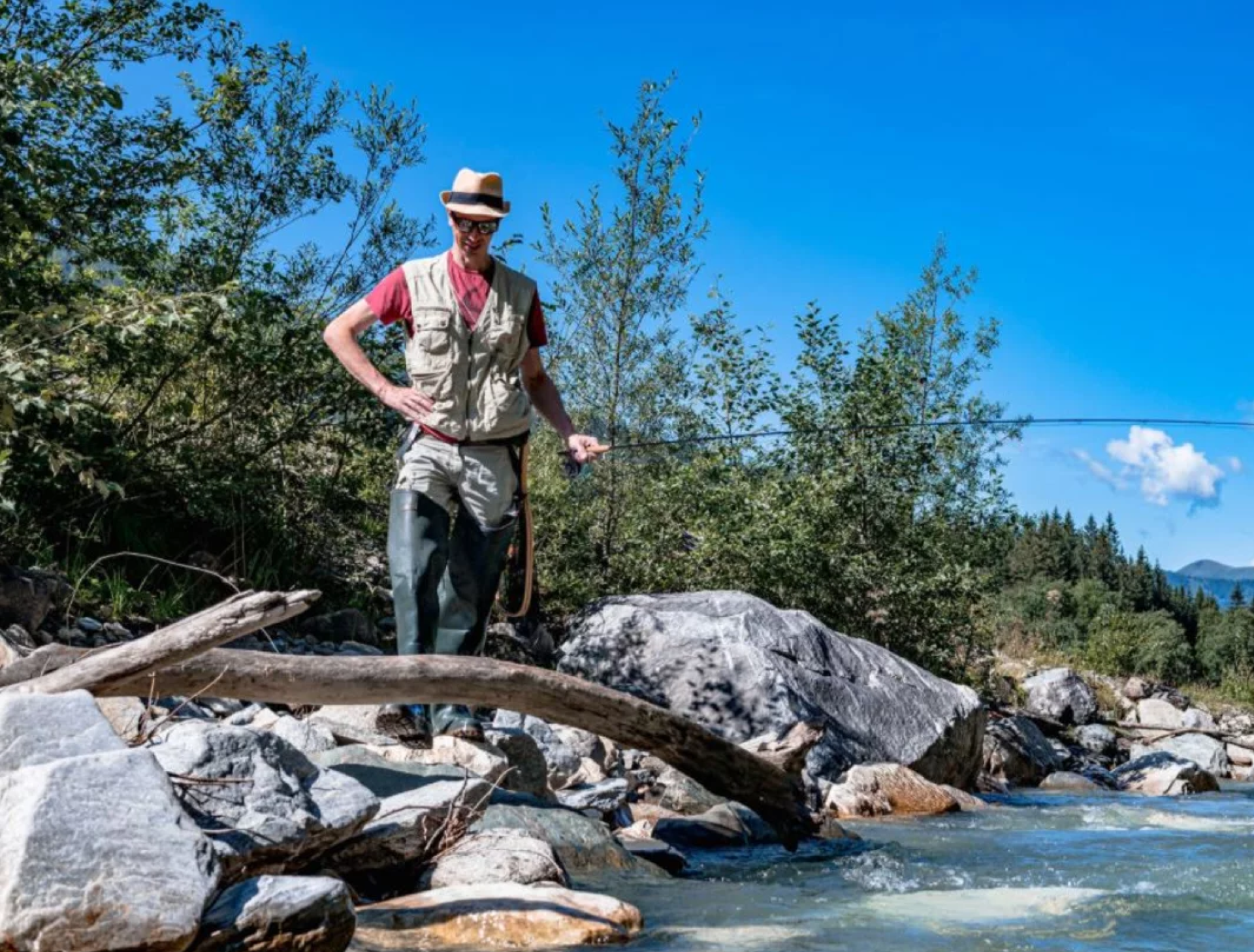 Eine Person mit Hut und Weste genießt das Fliegenfischen in einem malerischen, felsigen Fluss, umgeben von üppigem Grün und einem klaren blauen Himmel, der Ruhe vermittelt.