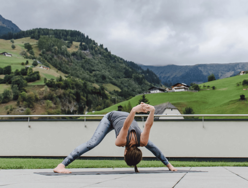 A woman practices yoga outdoors in a wide-legged forward bend pose, set against a backdrop of lush green hills and cloudy skies, conveying serenity.