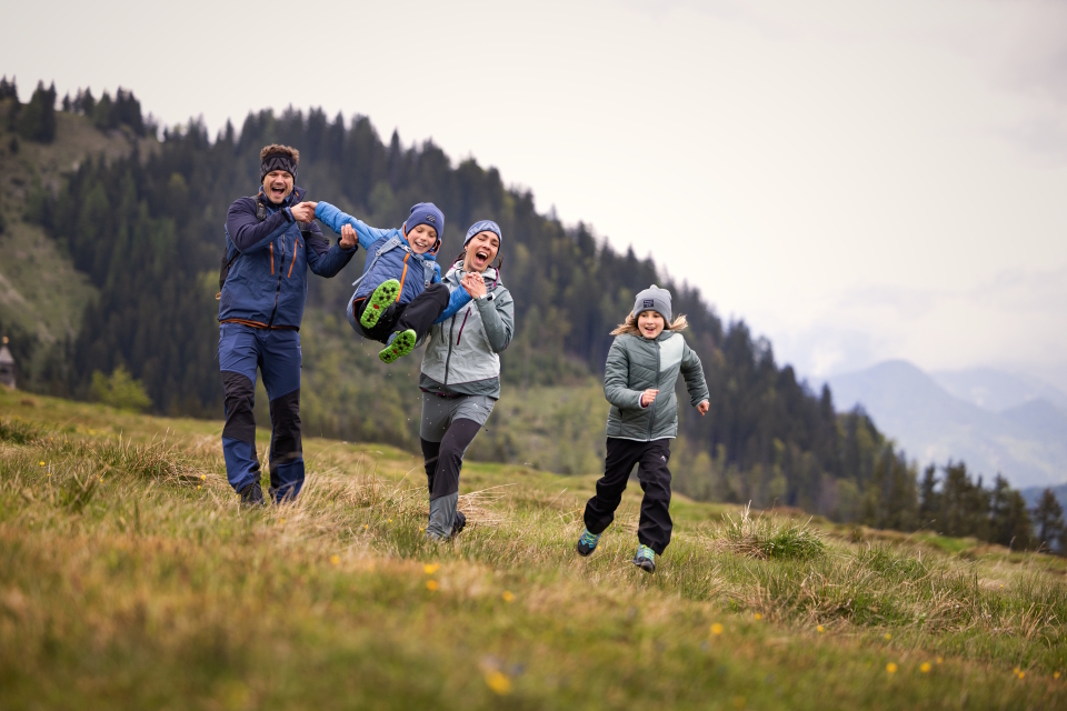 Eine fröhliche vierköpfige Familie rennt über eine grasbewachsene Wiese, umgeben von Bergen und Bäumen. Die Szene vermittelt Glück und Zusammengehörigkeit in der Natur.