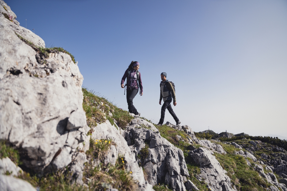 Zwei Wanderer in Outdoor-Kleidung wandern unter strahlend blauem Himmel auf einem felsigen Bergpfad. Die Landschaft ist zerklüftet und vermittelt ein Gefühl von Abenteuer und Entdeckungslust.