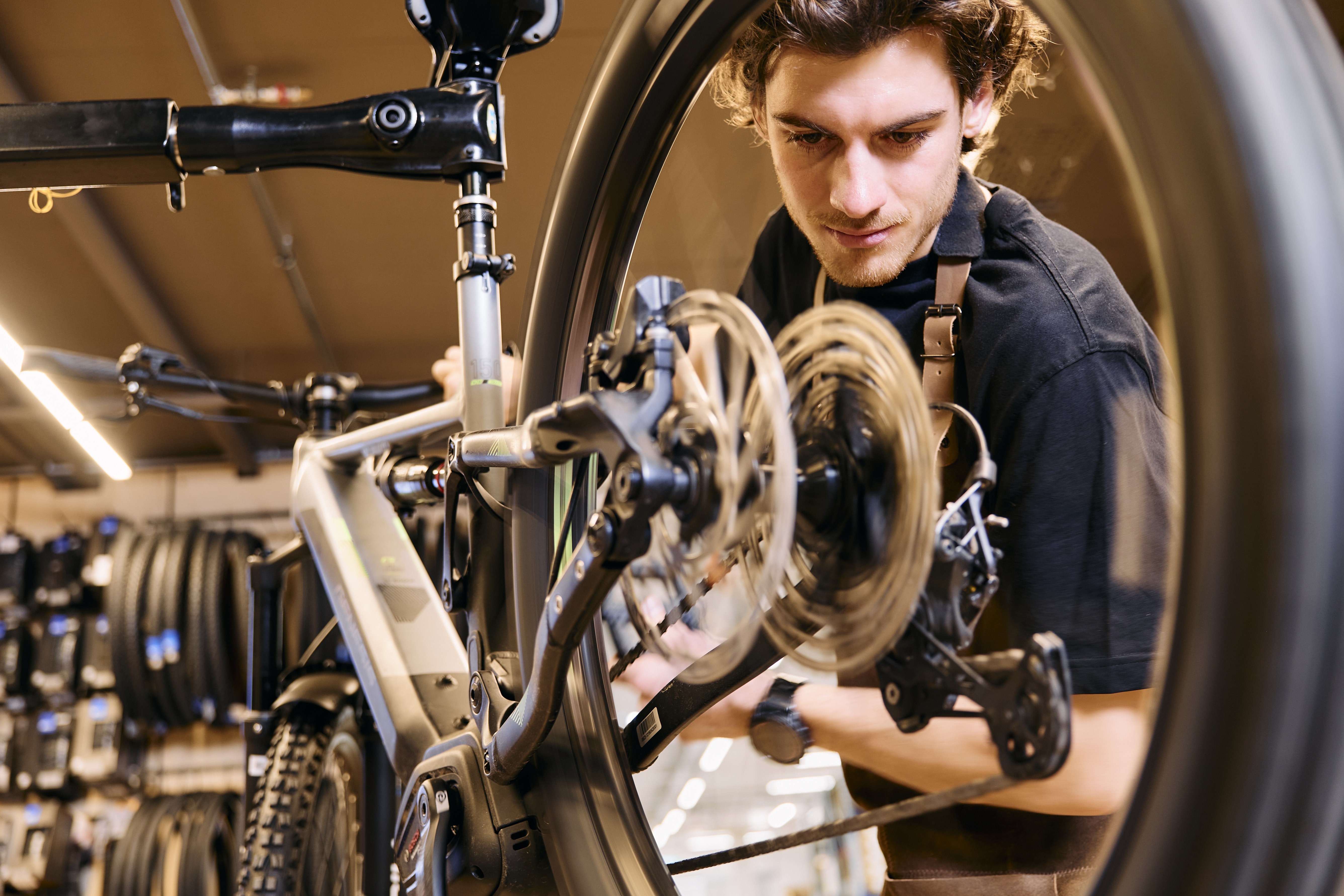 A focused mechanic in a workshop adjusts a mountain bike wheel. The background shows hanging tires, and the setting is warmly lit, creating an attentive mood.