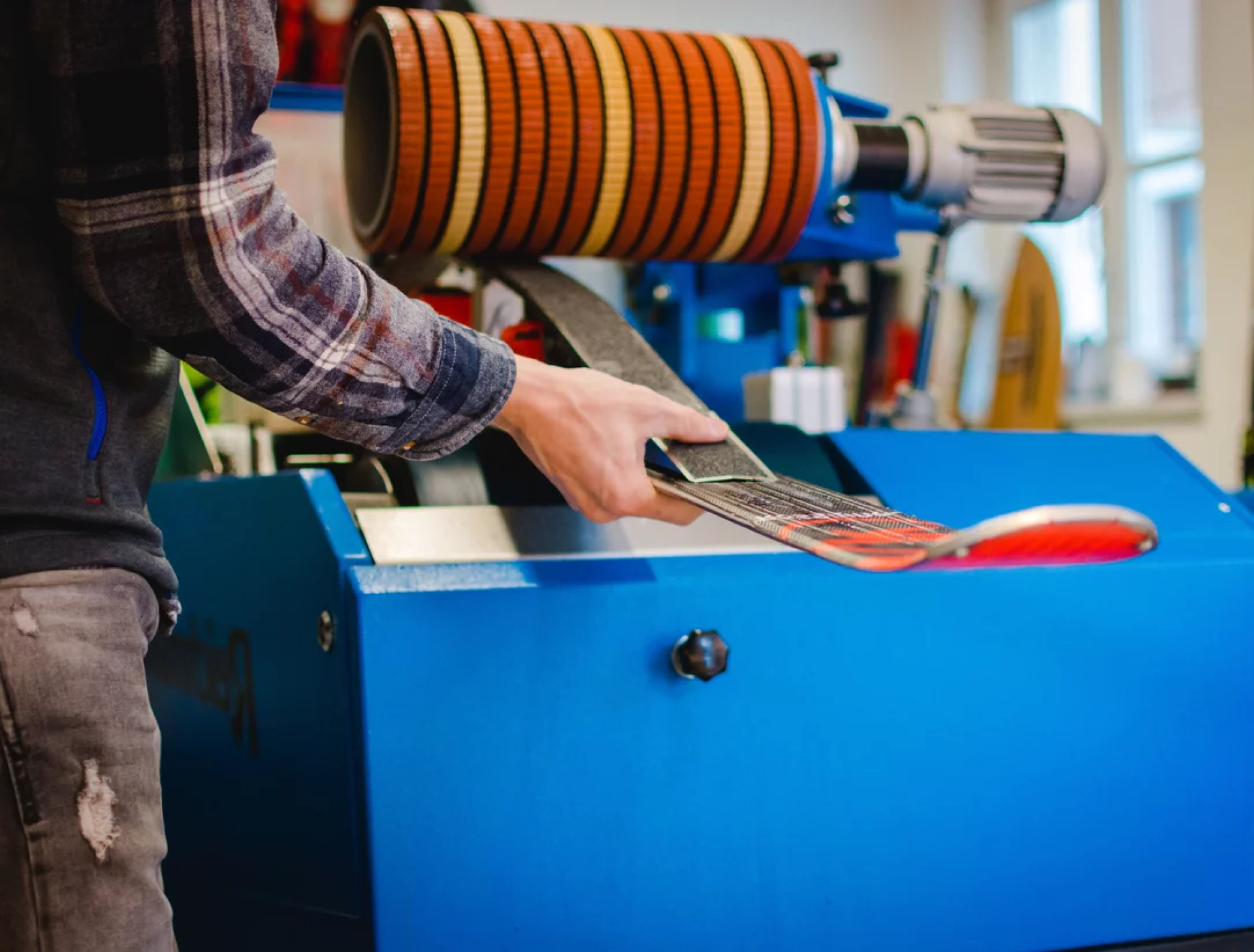 Eine Person stellt einen Ski auf einer blauen Schleifmaschine ein. Die Maschine verfügt über rotierende Schleifbänder. Die Einstellung wirkt technisch und konzentriert.