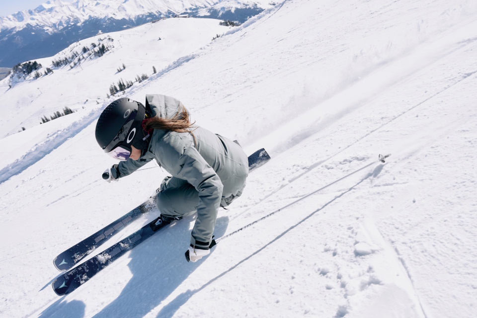 Ein Skifahrer in grauer Kleidung und Helm fährt gekonnt einen verschneiten Hang hinunter und wirbelt dabei Pulverschnee auf. Im Hintergrund sind schneebedeckte Berge und ein strahlend blauer Himmel zu sehen.