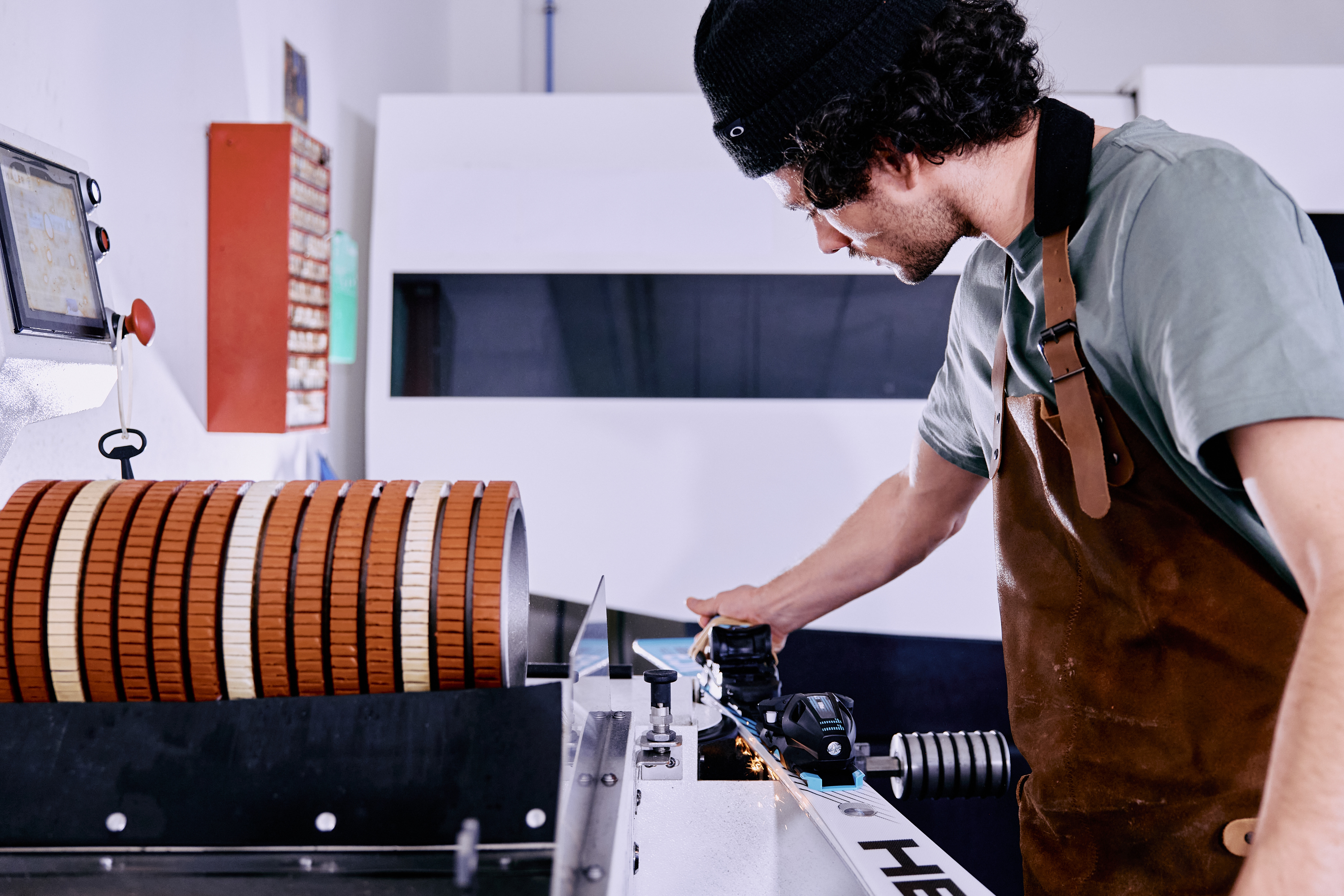 Eine Person mit schwarzer Mütze und brauner Schürze bedient in einer Werkstatt eine Maschine mit orangefarbenen Walzen und vermittelt dabei Konzentration und Präzision.