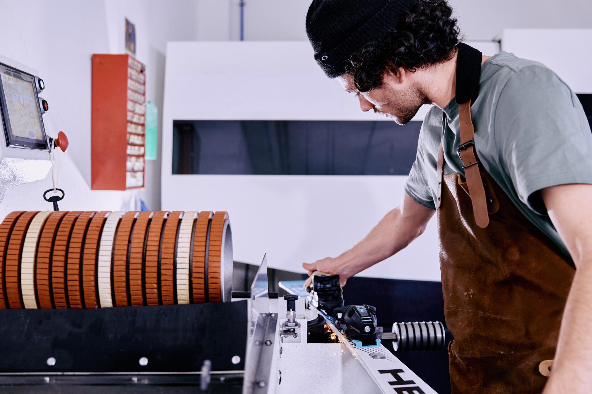 Eine Person mit schwarzer Mütze und brauner Schürze bedient in einer Werkstatt eine Maschine mit orangefarbenen Walzen und vermittelt dabei Konzentration und Präzision.