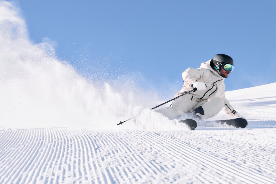 Ein Skifahrer in weißer Ausrüstung und Helm saust einen verschneiten Hang hinunter, lehnt sich in eine scharfe Kurve, über ihm strahlend blauer Himmel und hinter ihm eine Schneewolke.