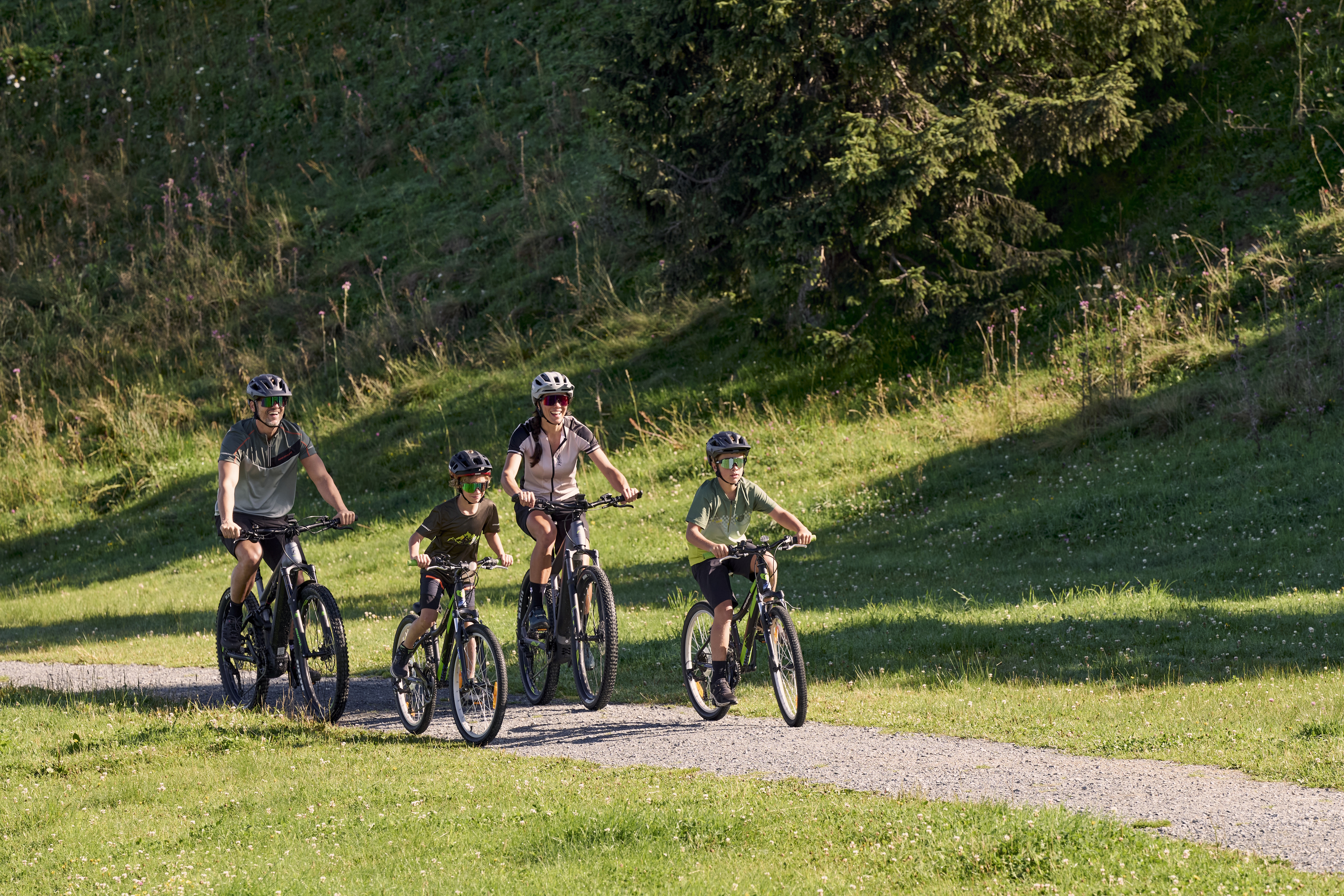 Eine vierköpfige Familie mit Helmen fährt an einem sonnigen Tag mit dem Fahrrad durch einen üppig grünen Park mit hohen Bäumen und Gras, was ein Gefühl von Glück und Zusammengehörigkeit vermittelt.