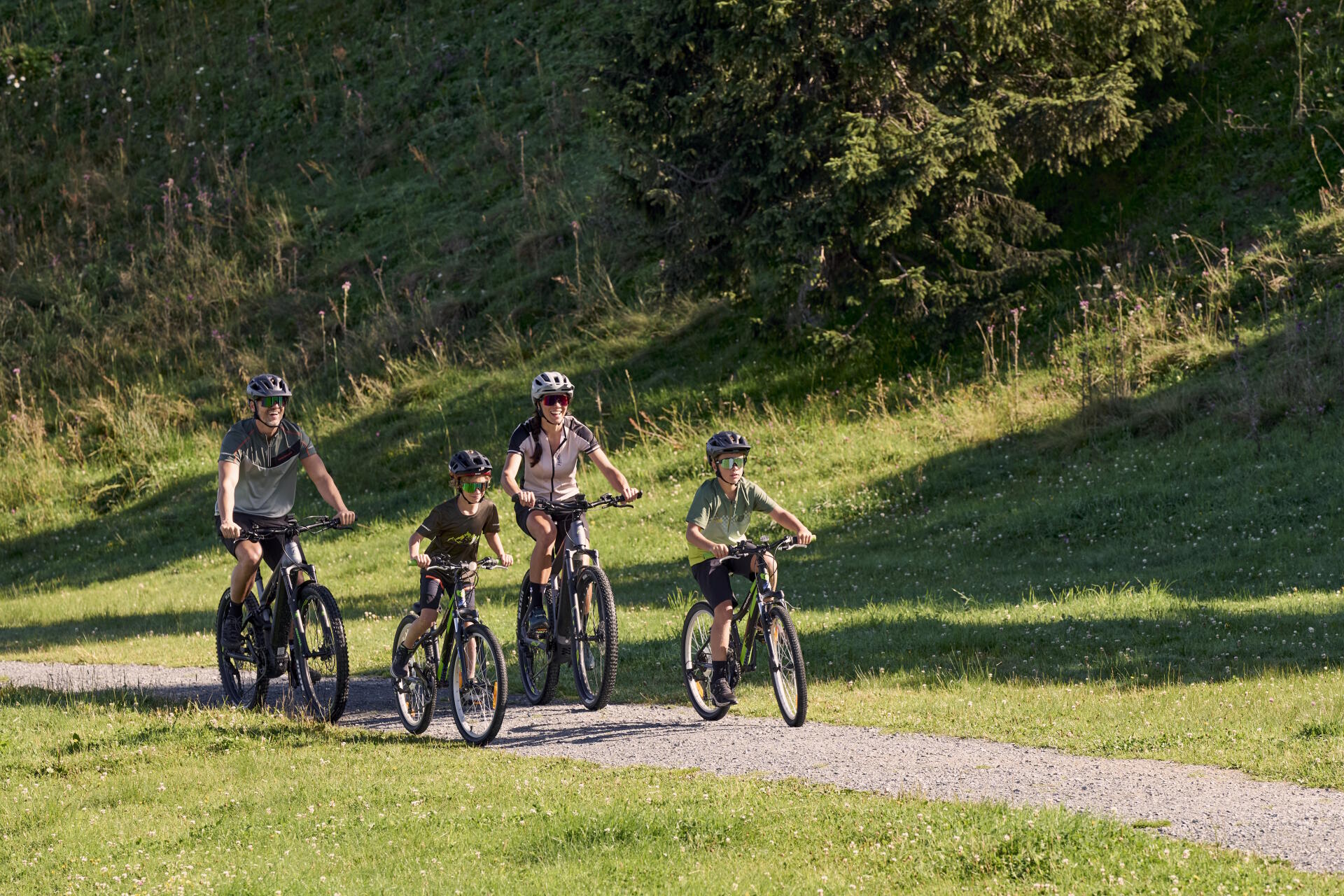 Eine vierköpfige Familie mit Helmen fährt an einem sonnigen Tag mit dem Fahrrad durch einen üppig grünen Park mit hohen Bäumen und Gras, was ein Gefühl von Glück und Zusammengehörigkeit vermittelt.