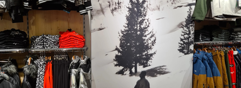 A store displays winter clothing, including jackets and pants, beside a monochrome mural of a snowy tree landscape, conveying a cozy, outdoor vibe.