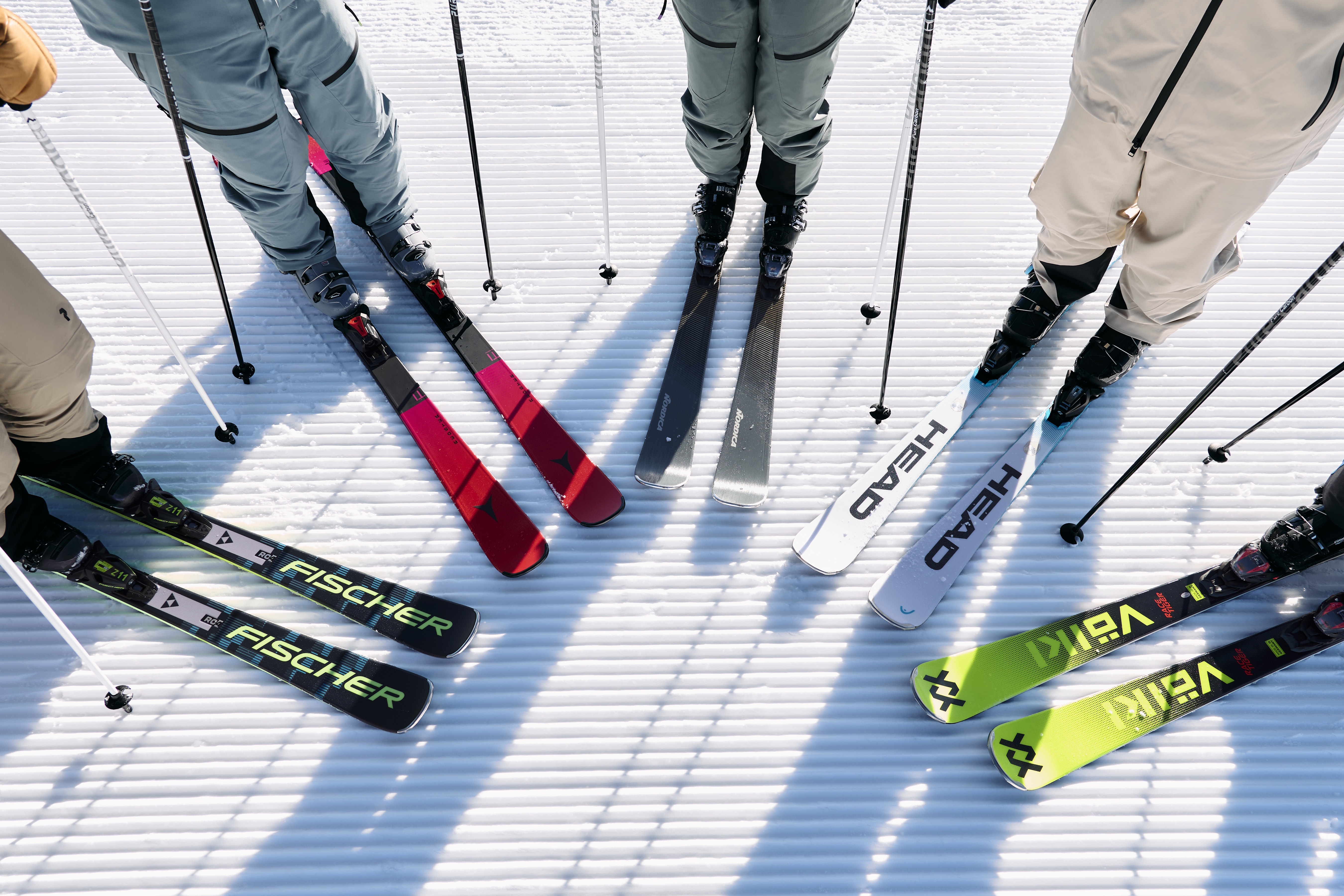 Four skiers stand on a groomed snow surface, ski tips converging. Skis display brand logos and vibrant colors, creating a dynamic, energetic scene.