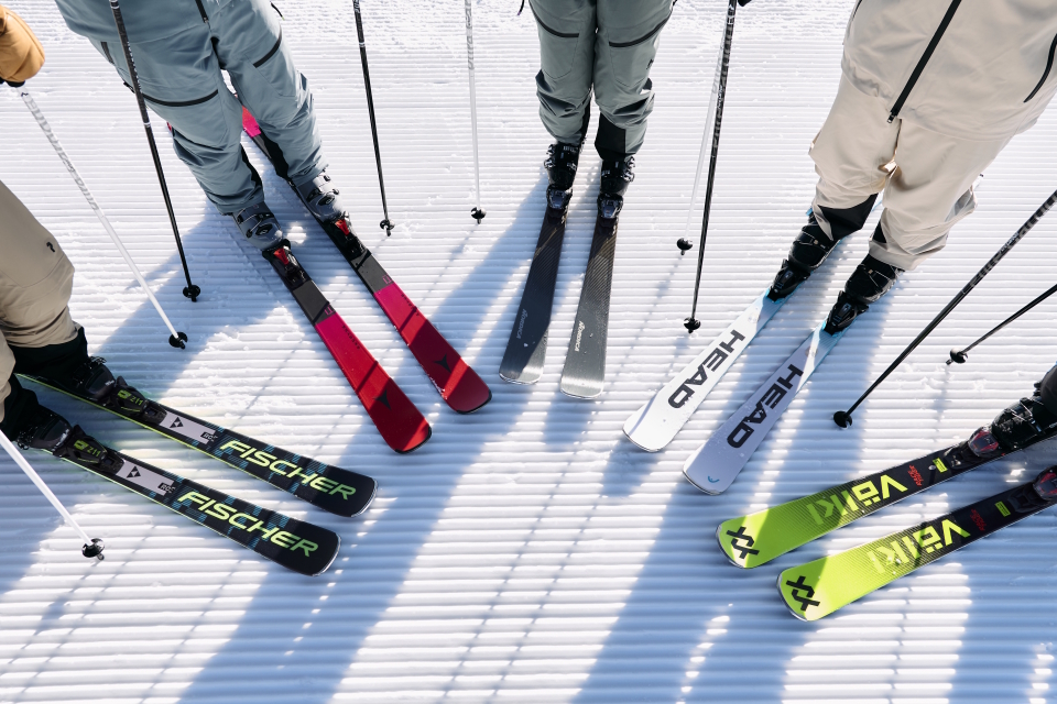 Vier Skifahrer stehen auf einer präparierten Schneefläche, die Skispitzen laufen zusammen. Die Skier sind mit Markenlogos und leuchtenden Farben versehen, wodurch eine dynamische, energiegeladene Szene entsteht.