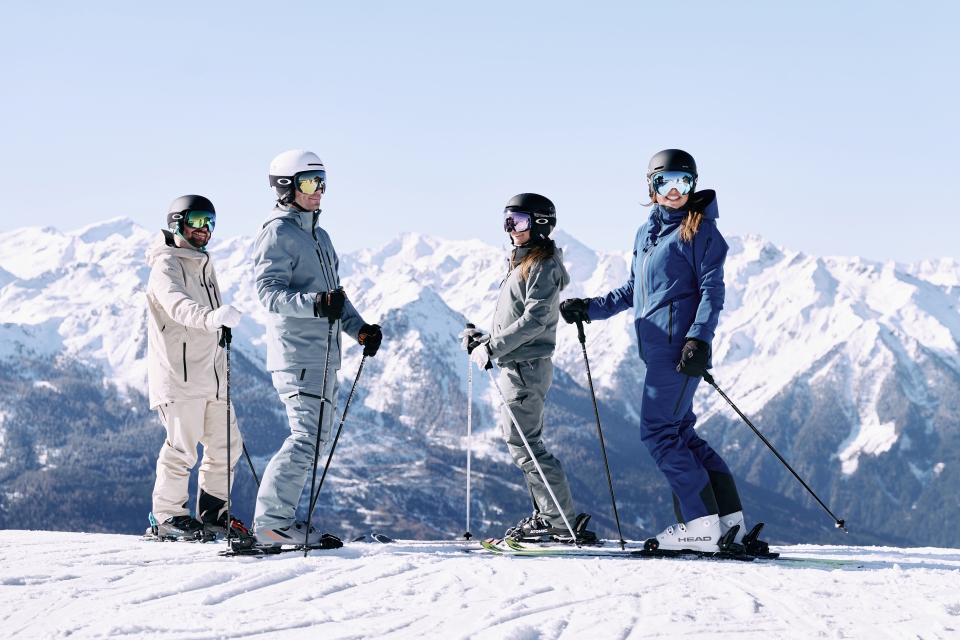 Vier Skifahrer in farbenfroher Ausrüstung stehen auf einem verschneiten Berghang, umgeben von majestätischen schneebedeckten Gipfeln unter einem strahlend blauen Himmel, und vermitteln Freude und Abenteuerlust.