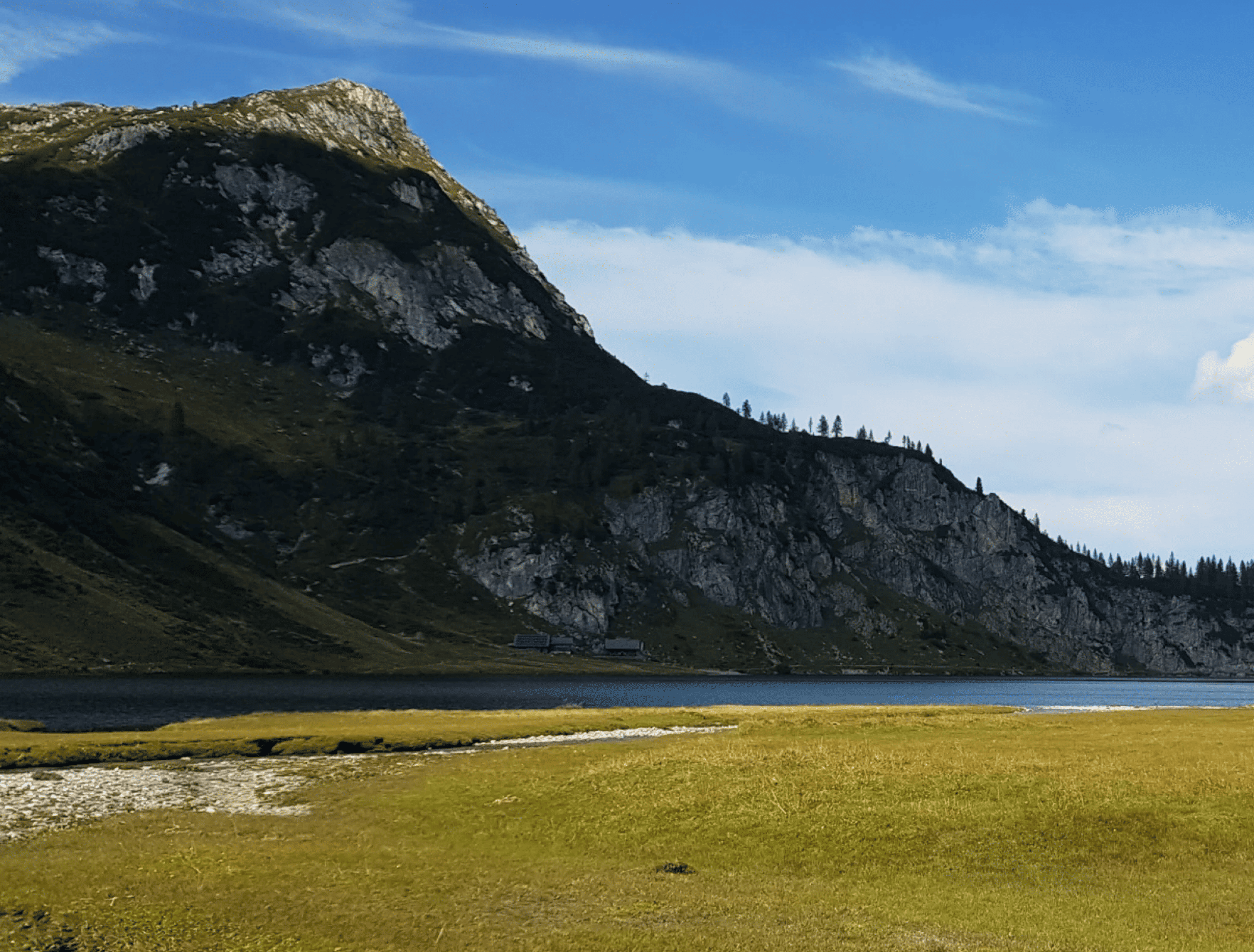 Eine ruhige Landschaft mit einer Wiese im Vordergrund, einem stillen See und einem zerklüfteten Berg unter einem klaren blauen Himmel, die Gelassenheit vermittelt.