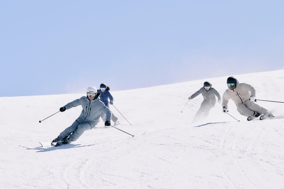 Vier Skifahrer in Winterausrüstung gleiten vor einem strahlend blauen Himmel einen verschneiten Hang hinunter und strahlen dabei Begeisterung und Können aus, während sie sich dynamisch und koordiniert die Abfahrt hinunterbewegen.
