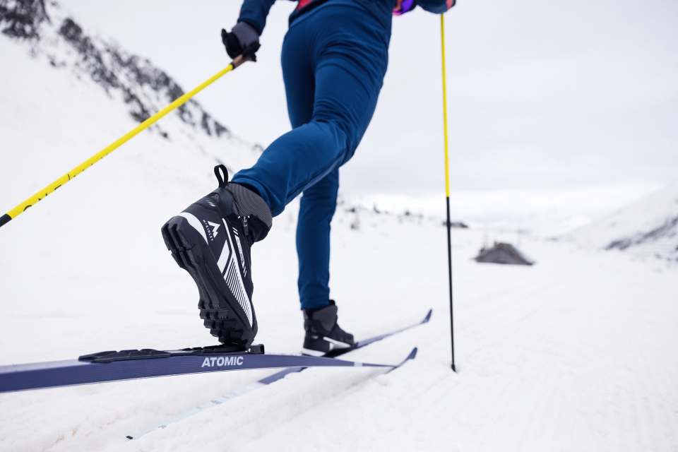 Person beim Langlaufen in einer verschneiten Landschaft, bekleidet mit blauer Skiausrüstung. Fokus auf Skischuhe und Skistöcke mit schneebedeckten Bergen im Hintergrund. Dynamische Stimmung.