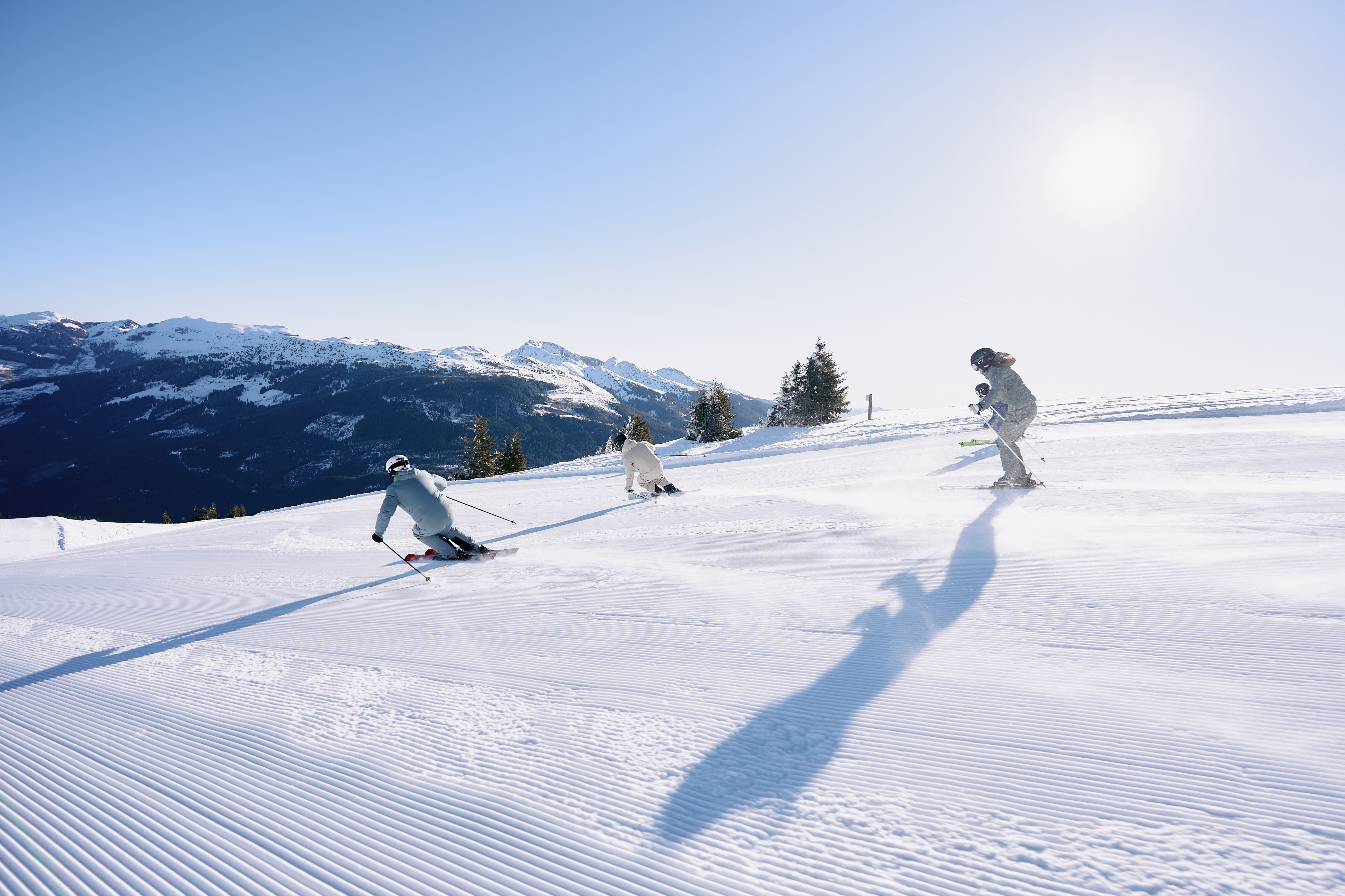 Three skiers glide down a sunlit, snow-covered slope with mountainous landscape in the background. Clear blue sky and long shadows convey a serene winter scene.
