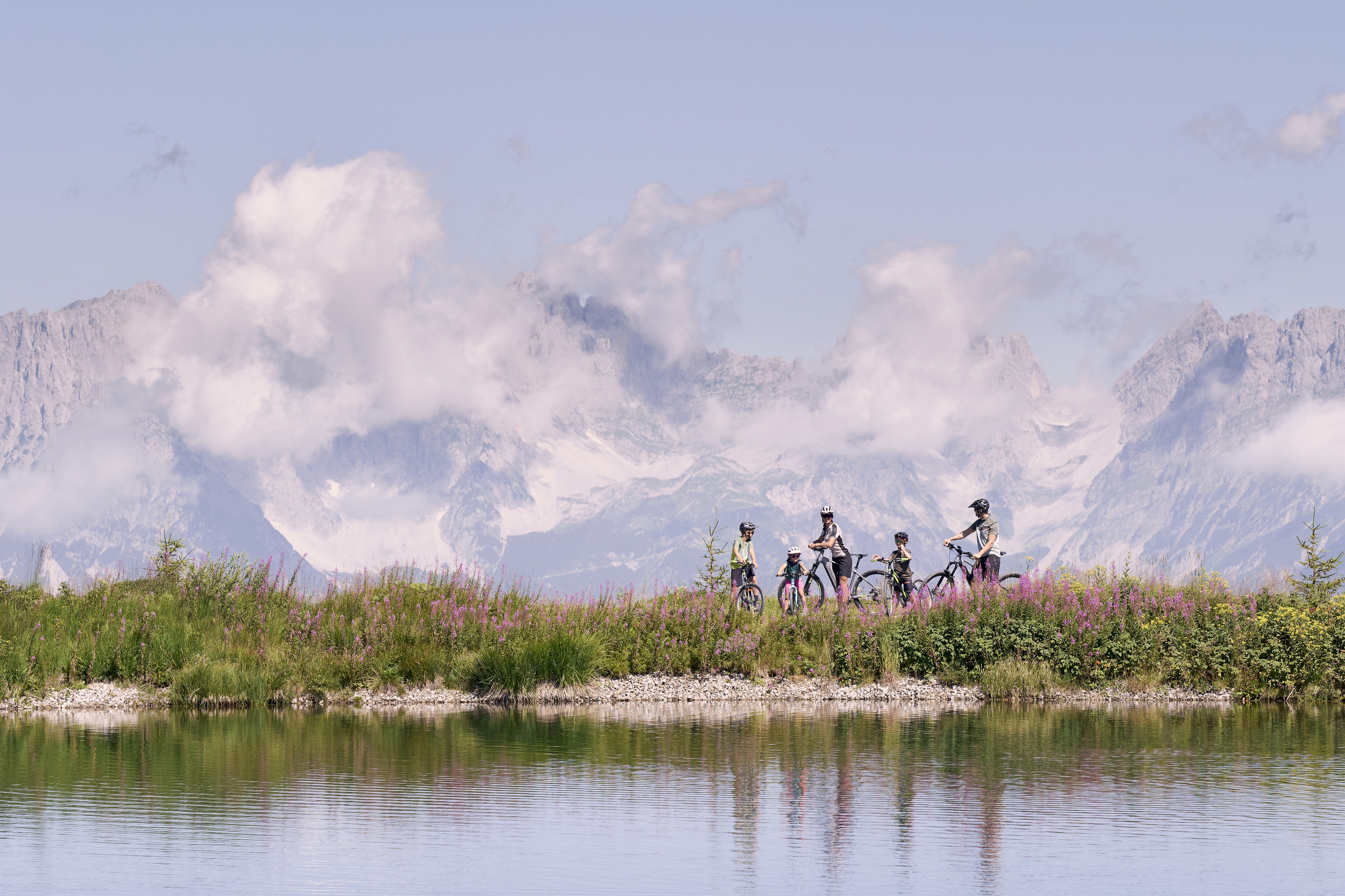 A group of five cyclists ride along a grassy path by a tranquil lake, with majestic, cloud-covered mountains in the background, conveying adventure and serenity.