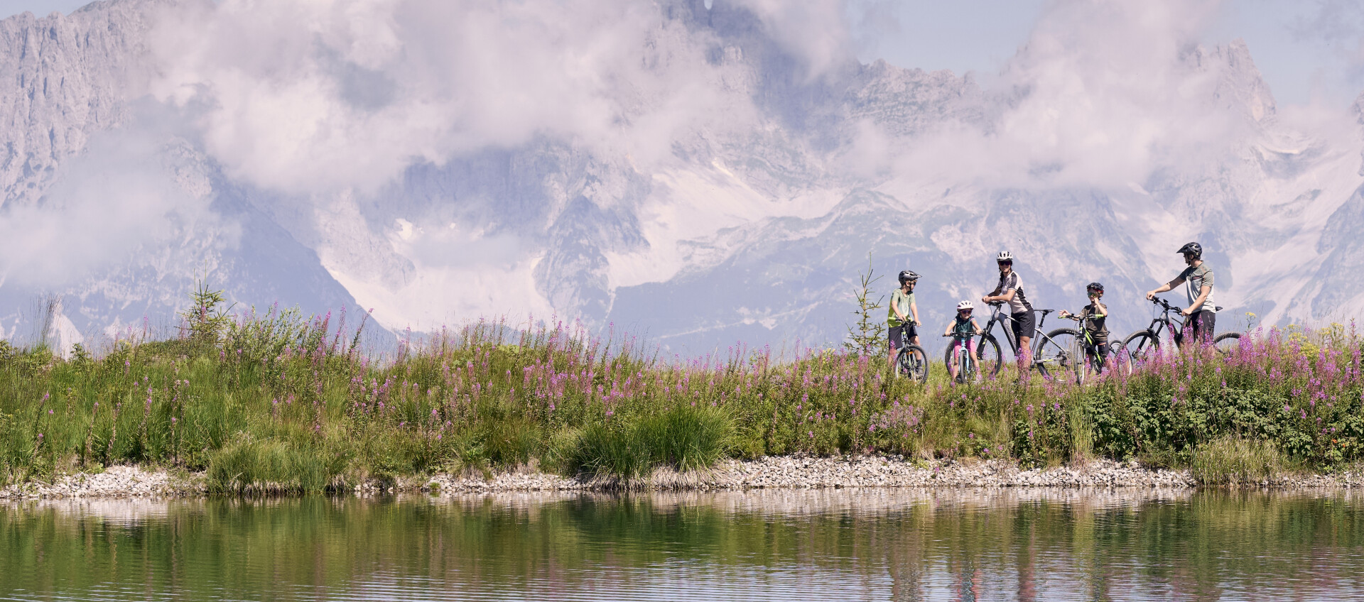 Gruppenradtour an einem malerischen Bergsee, Fahrräder an einem Wildblumenhang, mit bewölkten Gipfeln in der Ferne.