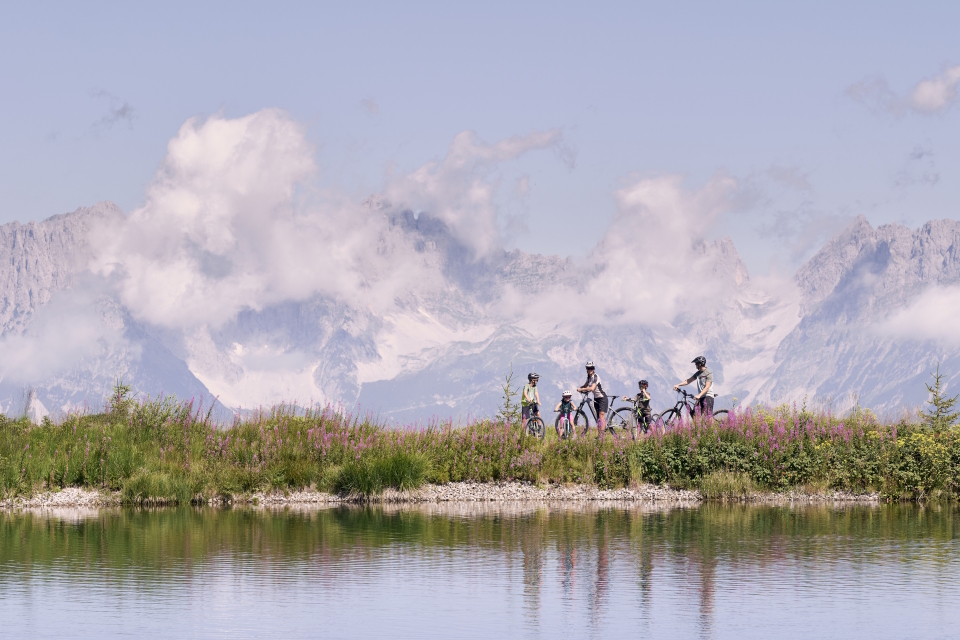 Eine Gruppe von fünf Radfahrern fährt auf einem grasbewachsenen Weg an einem ruhigen See entlang, im Hintergrund majestätische, wolkenverhangene Berge, die Abenteuer und Gelassenheit vermitteln.