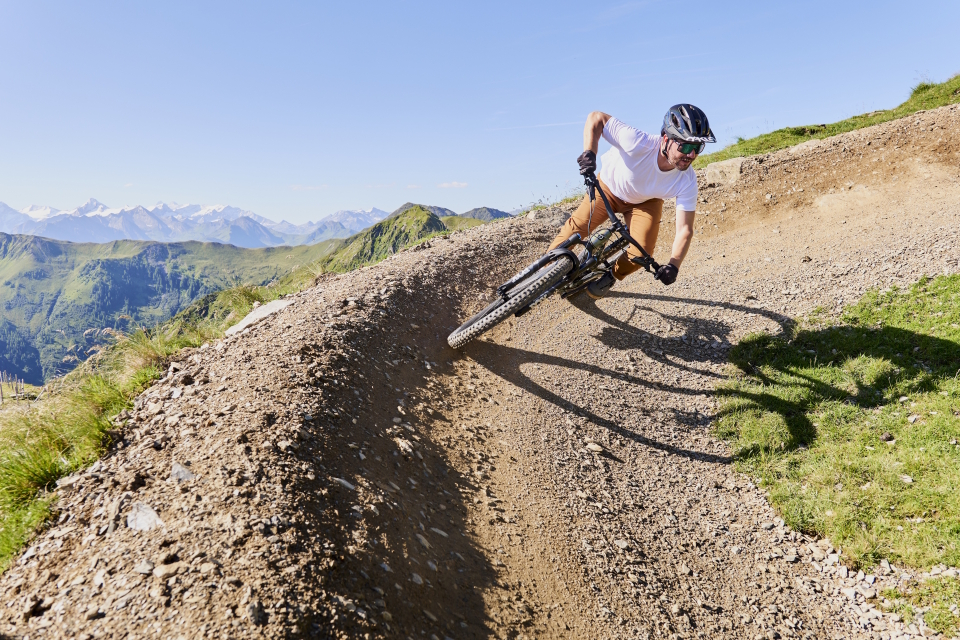 A cyclist leans into a curve on a mountain trail, surrounded by green hills and distant snowy peaks under a clear blue sky, conveying a sense of adventure.