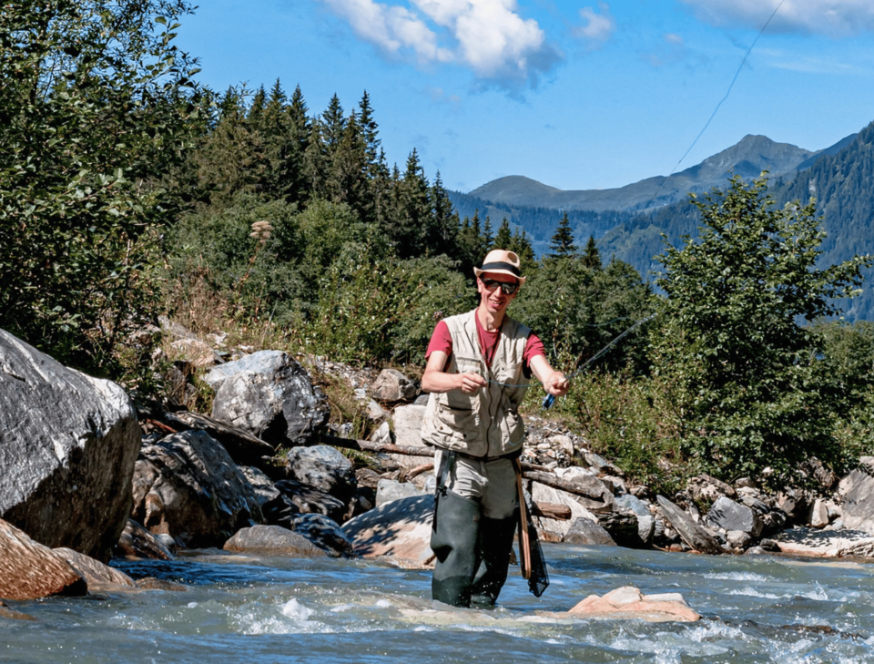 A person fly-fishing in a rocky mountain stream, surrounded by lush trees and distant peaks under a blue sky, conveying a sense of tranquility.