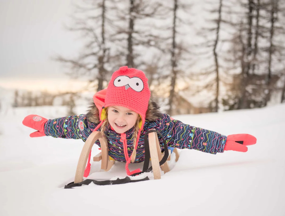 Ein fröhliches Kind in einer bunten Jacke und einer roten Mütze fährt mit ausgestreckten Armen auf einem Schlitten über den Schnee. Im Hintergrund stehen kahle Bäume vor einem sanften Himmel.